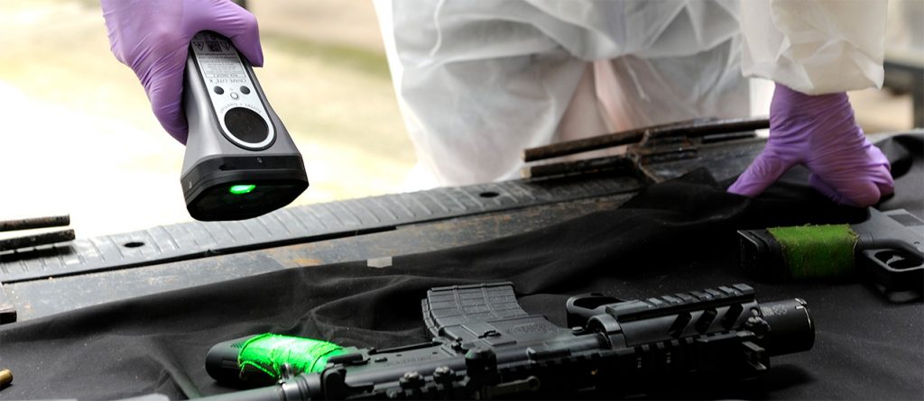 Forensic examiner using the Crime-Lite X to identify evidence on the side of a gun.